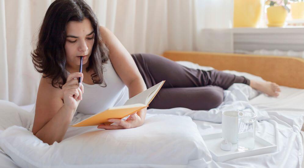 A person sitting at a kitchen table in the morning light, writing in a small notebook next to a mug and a phone placed face down